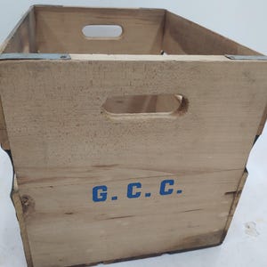 Op de afbeelding: Een vintage houten krat met uitgesneden handgrepen en metalen verstevigingen. De krat is gemaakt van lichtgekleurd hout en heeft de blauwe letters "G.C.C." op de zijkant. De krat is leeg en lijkt goed gebruikt.