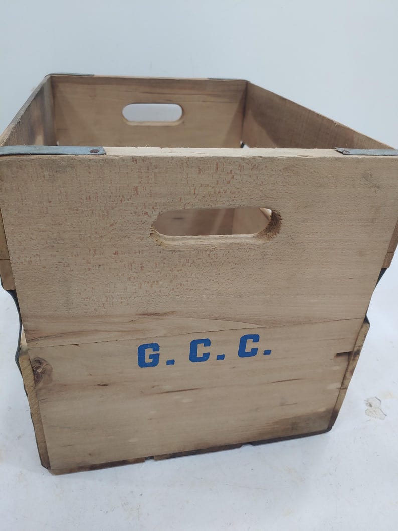 Op de afbeelding: Een vintage houten krat met uitgesneden handgrepen en metalen verstevigingen. De krat is gemaakt van lichtgekleurd hout en heeft de blauwe letters "G.C.C." op de zijkant. De krat is leeg en lijkt goed gebruikt.