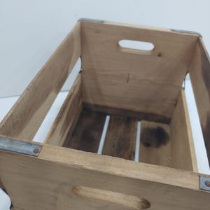 Op de afbeelding: Houten krat met metalen hoekverstevigingen en handgrepen. De krat is gemaakt van lichtgekleurd hout met zichtbare nerven en wat donkere vlekken. De letters "G.C.C." zijn blauw op de voorkant gestencild.
