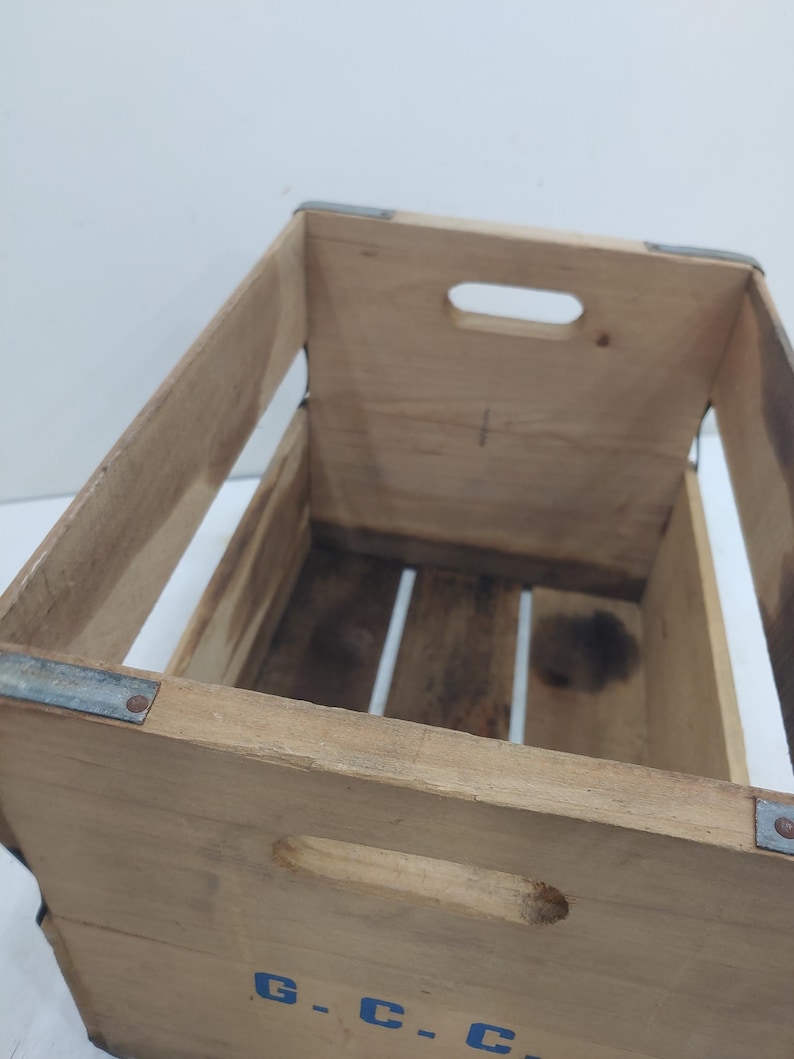 Op de afbeelding: Houten krat met metalen hoekverstevigingen en handgrepen. De krat is gemaakt van lichtgekleurd hout met zichtbare nerven en wat donkere vlekken. De letters "G.C.C." zijn blauw op de voorkant gestencild.
