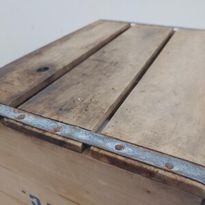 Op de afbeelding: Een vintage houten krat met een lichtbruine afwerking. De bovenkant is gemaakt van parallelle houten latten en de hoeken zijn verstevigd met metalen banden die met klinknagels zijn bevestigd. Het hout vertoont tekenen van slijtage en ouderdom.