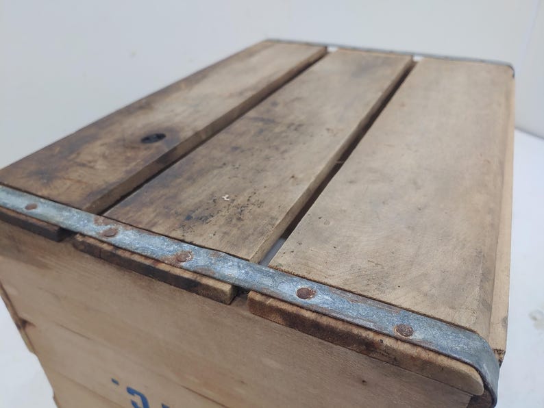 Op de afbeelding: Een vintage houten krat met een lichtbruine afwerking. De bovenkant is gemaakt van parallelle houten latten en de hoeken zijn verstevigd met metalen banden die met klinknagels zijn bevestigd. Het hout vertoont tekenen van slijtage en ouderdom.