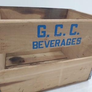 Op de afbeelding: Vintage houten drankkrat met blauwe letters "G.C.C. BEVERAGES". De krat heeft metalen verstevigingen op de hoeken en randen. Het hout heeft een natuurlijke, verouderde uitstraling.