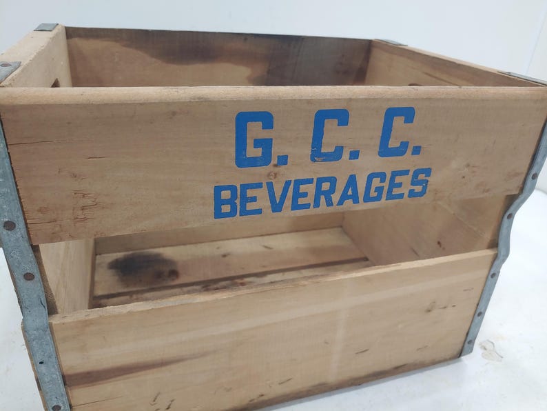 Op de afbeelding: Vintage houten drankkrat met blauwe letters "G.C.C. BEVERAGES". De krat heeft metalen verstevigingen op de hoeken en randen. Het hout heeft een natuurlijke, verouderde uitstraling.