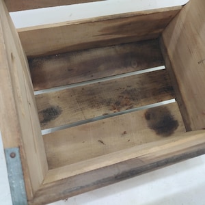 Op de afbeelding: Een houten krat met een rechthoekige vorm, met een lattenbodem en zijkanten. De hoeken zijn versterkt met metaal. Het hout heeft een natuurlijke, verweerde uitstraling met donkere vlekken. Er is een handgreepopening aan &eacute;&eacute;n kant.