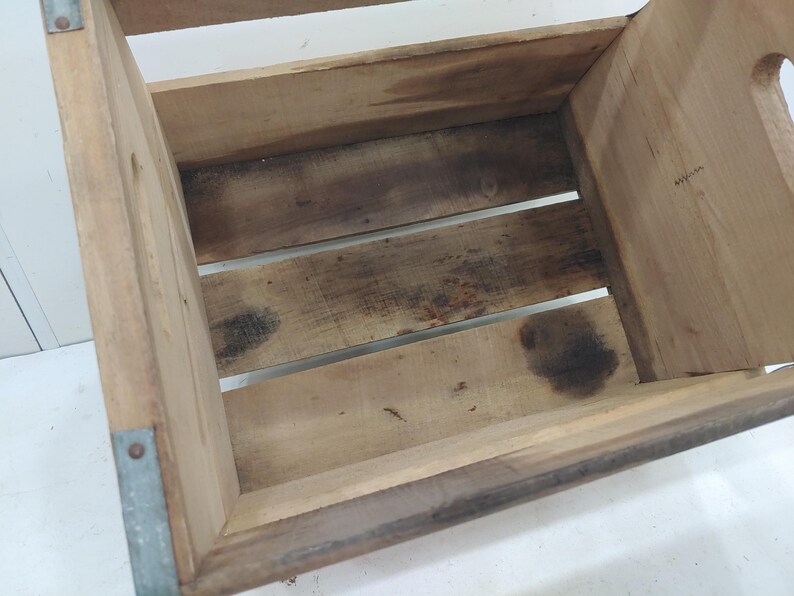 Op de afbeelding: Een houten krat met een rechthoekige vorm, met een lattenbodem en zijkanten. De hoeken zijn versterkt met metaal. Het hout heeft een natuurlijke, verweerde uitstraling met donkere vlekken. Er is een handgreepopening aan &eacute;&eacute;n kant.