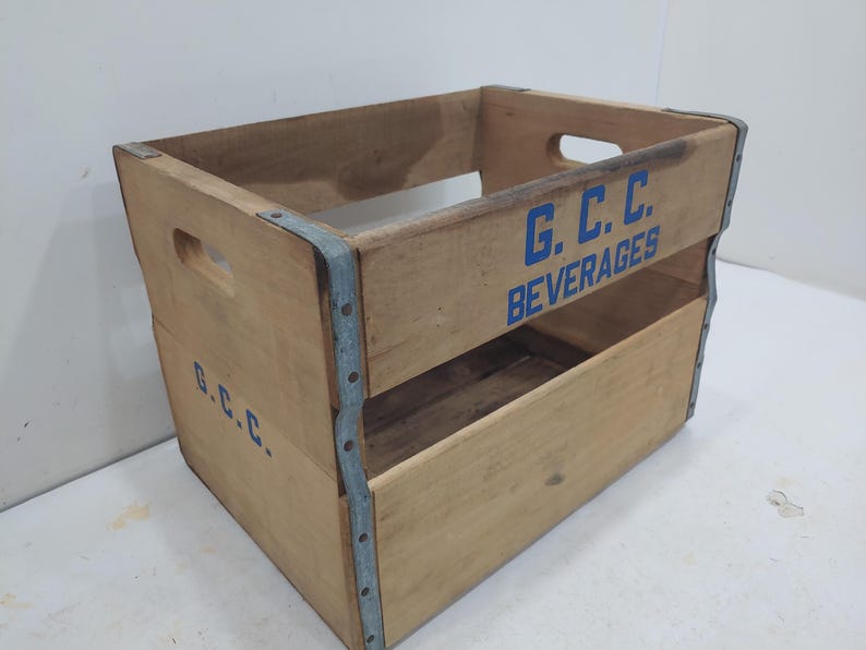 Op de afbeelding: Een vintage houten drankkrat met de tekst "G.C.C. BEVERAGES" in blauwe letters. De krat heeft een rechthoekige vorm met handgesneden openingen aan de zijkanten om te dragen. Metalen strips versterken de hoeken en randen, wat bijdraagt aan de rustieke charme.