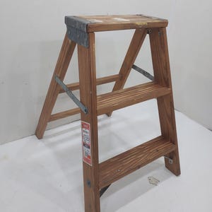 Könnte beinhalten: Ein Holztritt mit drei Stufen. Der Tritt besteht aus hellbraunem Holz und hat Metallwinkel an den Ecken und Seiten zur Verstärkung. Ein rot-weißes Etikett ist an einem der Beine angebracht.