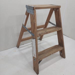 Könnte beinhalten: Eine Holztrittleiter mit drei Stufen und einer oberen Plattform. Die Leiter besteht aus hellfarbenem Holz mit Metallwinkeln und -stützen. Die Farbe ist stellenweise abgeblättert, was ihr einen Vintage-Look verleiht.