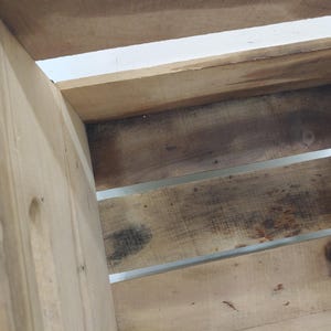 Op de afbeelding: Close-up van een houten krat met zichtbare houtnerf en metalen accenten. De krat is gemaakt van lichtgekleurd hout met donkere, verouderde vlekken. Het ontwerp heeft horizontale latten en een stevige, rustieke uitstraling.