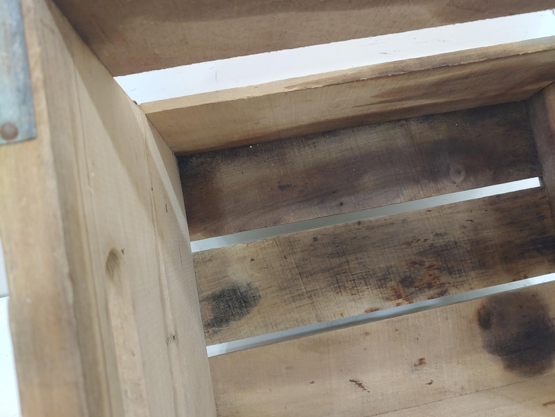 Op de afbeelding: Close-up van een houten krat met zichtbare houtnerf en metalen accenten. De krat is gemaakt van lichtgekleurd hout met donkere, verouderde vlekken. Het ontwerp heeft horizontale latten en een stevige, rustieke uitstraling.