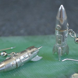 May include: A pair of silver rocket ship earrings. The earrings are made of metal and have a detailed design. The rocket ships are about 1 inch long and have a silver hook.