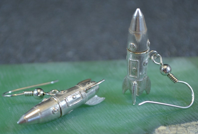 May include: A pair of silver rocket ship earrings. The earrings are made of metal and have a detailed design. The rocket ships are about 1 inch long and have a silver hook.