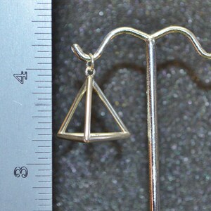 Puede incluir: Encanto de colgante de pir&aacute;mide geom&eacute;trica de plata colgado de un soporte de exhibici&oacute;n. El colgante es una pir&aacute;mide hueca tridimensional con un marco de alambre.