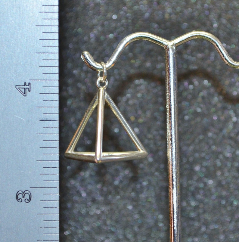 Puede incluir: Encanto de colgante de pir&aacute;mide geom&eacute;trica de plata colgado de un soporte de exhibici&oacute;n. El colgante es una pir&aacute;mide hueca tridimensional con un marco de alambre.