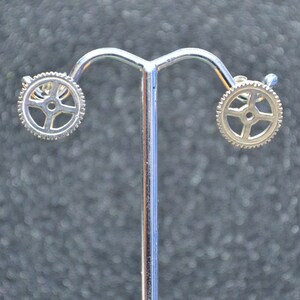 May include: A pair of silver earrings with a gear design. The earrings are displayed on a silver stand.