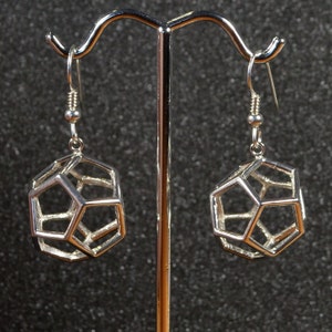 Puede incluir: Un par de pendientes colgantes geométricos plateados con un diseño en forma de jaula. Los pendientes están colgados de un soporte plateado.