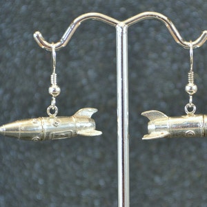 May include: A pair of silver rocket ship earrings. The earrings are hanging from a silver stand.