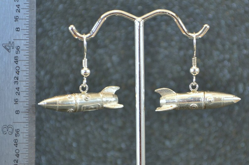 May include: A pair of silver rocket ship earrings. The earrings are hanging from a silver stand.