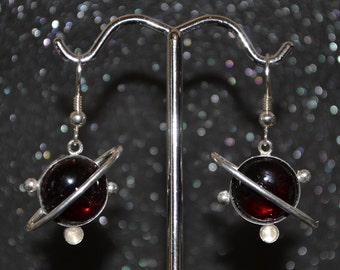 May include: A pair of silver earrings with a red glass orb and a silver ring around it, resembling a planet.