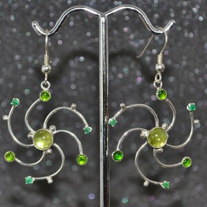 May include: Silver earrings with a swirling design and green gemstones. The earrings feature a larger green gemstone in the center of each earring.