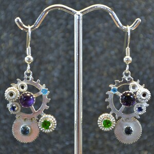 Peut inclure: Boucles d'oreilles en argent avec un design steampunk comprenant des engrenages, des pierres précieuses violettes, bleues et vertes.