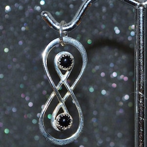 Peut inclure: Un pendentif en argent en forme de symbole de l'infini avec deux pierres précieuses noires. Le pendentif est suspendu à un crochet en argent.