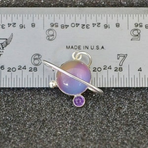 May include: A silver charm in the shape of a planet with a purple and blue iridescent glass orb and a silver ring around it. The charm has a small purple gemstone on the bottom.