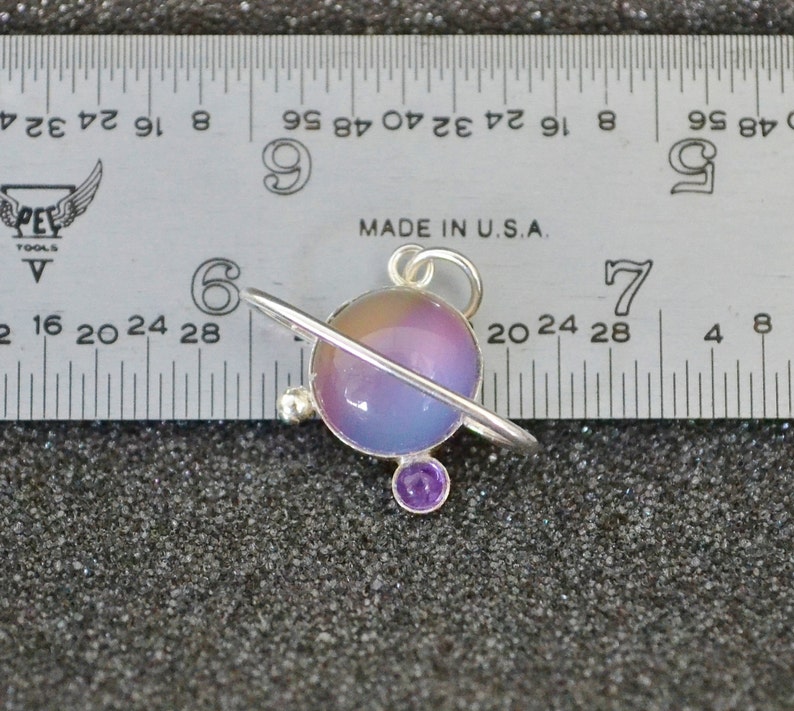 May include: A silver charm in the shape of a planet with a purple and blue iridescent glass orb and a silver ring around it. The charm has a small purple gemstone on the bottom.