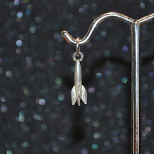 May include: A silver rocket ship charm pendant hanging from a silver display stand against a black background with out-of-focus silver glitter.