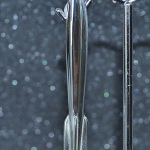 May include: A shiny, silver-colored rocket-shaped object. The rocket has a pointed tip and a detailed base with vertical lines. It is displayed on a silver metal stand against a dark, bokeh background.