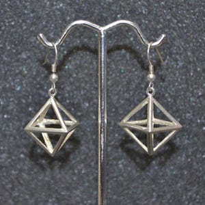 Puede incluir: Pendientes geométricos plateados con forma de diamante tridimensional. Los pendientes se exhiben en un soporte plateado sobre un fondo oscuro y moteado. Cierre de gancho.