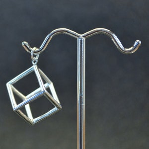 Puede incluir: Un colgante geométrico plateado en forma de cubo con un centro hueco, colgado de una cadena plateada en un expositor.