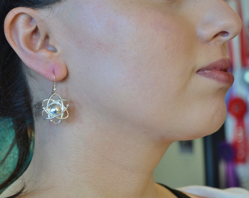 Pu&ograve; includere: Orecchini in argento a forma di atomo con una sfera rotonda in argento al centro. Gli orecchini sono appesi all'orecchio di una persona.