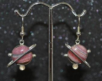 May include: A pair of silver earrings with pink and white striped gemstone spheres and silver rings around them, resembling planets.