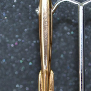 May include: A gold-colored metal rocket-shaped pin. The pin is about 2 inches long and has a pointed tip. The pin is on a display stand.
