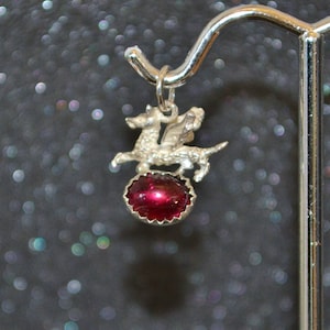 May include: Silver charm featuring a winged dragon perched atop a red gemstone.