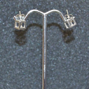 Może przedstawiać: Para srebrnych kolczyków w kształcie sześcianu z pustym środkiem. Kolczyki są prezentowane na srebrnym stojaku.