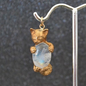 Könnte beinhalten: Ein goldfarbener Katzenanhänger, der einen ovalen, blau-weißen Edelstein hält. Die Katze hat geschlossene Augen und umklammert den Stein mit ihren Pfoten. Der Anhänger ist an einer goldfarbenen Öse aufgehängt.