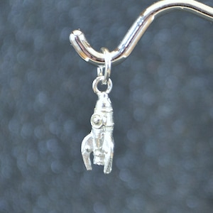 Puede incluir: Un amuleto de plata con forma de cohete. El amuleto cuelga de un gancho de plata.