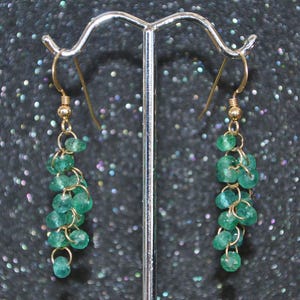 May include: A pair of gold-colored earrings featuring cascading clusters of small, faceted emerald green beads. The earrings hang from gold-colored ear wires, creating a delicate and elegant design. The beads are strung together with gold-colored wire.
