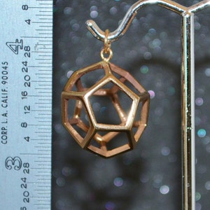 Pode incluir: Um pendente geom&eacute;trico dourado com um design de dodecaedro, suspenso por um gancho dourado. O pendente tem aproximadamente 2,5 cm de di&acirc;metro. Uma r&eacute;gua com marca&ccedil;&otilde;es em cm &eacute; vis&iacute;vel no fundo.