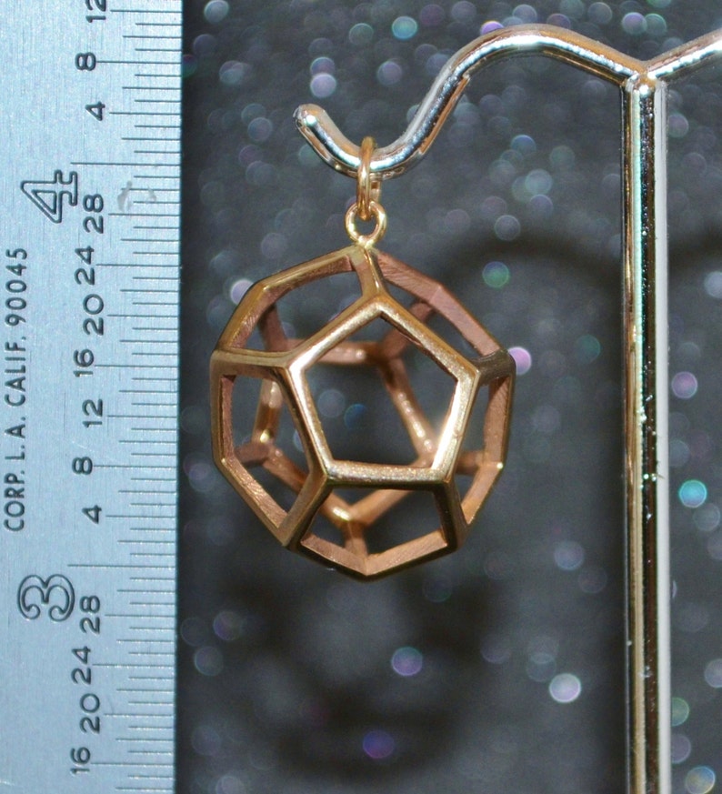 Pode incluir: Um pendente geom&eacute;trico dourado com um design de dodecaedro, suspenso por um gancho dourado. O pendente tem aproximadamente 2,5 cm de di&acirc;metro. Uma r&eacute;gua com marca&ccedil;&otilde;es em cm &eacute; vis&iacute;vel no fundo.