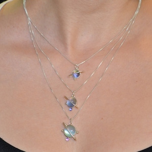 May include: A silver necklace with three layered chains and three planet-shaped pendants. The pendants are made of iridescent glass and have a silver ring around them. The smallest pendant is at the bottom of the necklace, and the largest pendant is at the top.