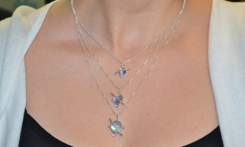 May include: A silver necklace with three layered chains and three planet-shaped pendants. The pendants are made of iridescent glass and have a silver ring around them. The smallest pendant is at the bottom of the necklace, and the largest pendant is at the top.