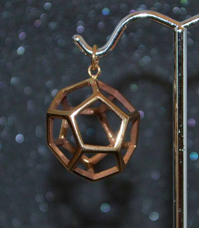 Pode incluir: Um pendente geom&eacute;trico em forma de dodecaedro, feito de metal dourado. O design vazado revela a estrutura interna. O pendente est&aacute; pendurado em um pequeno la&ccedil;o dourado, contra um fundo escuro e desfocado.