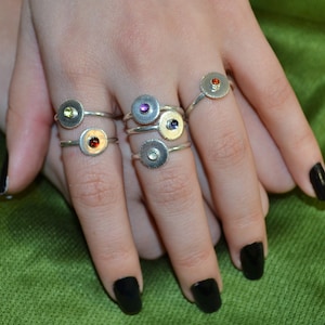 May include: A hand model wearing five silver rings with colourful gemstones. Each ring features a round, textured band with a small, circular gemstone in the centre. The gemstones are purple, yellow, red, green, and blue.