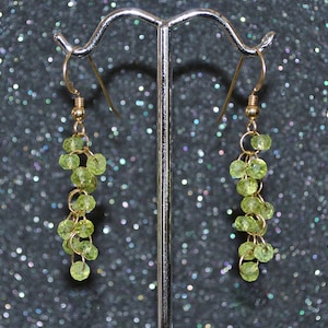 May include: A pair of gold dangle earrings with green gemstone beads. The earrings are hanging from a silver stand.
