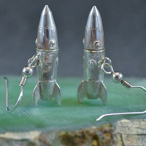 May include: A pair of silver rocket ship earrings with a dangling hook closure. The rockets have a textured surface and a silver finish.