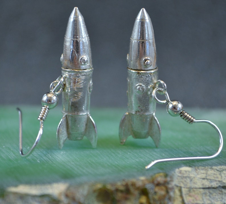 May include: A pair of silver rocket ship earrings with a dangling hook closure. The rockets have a textured surface and a silver finish.